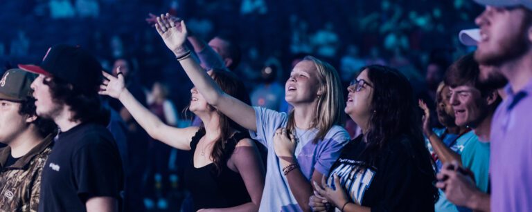colorful photo of the audience in a dark auditorium worshiping with eyes closed and hands extended