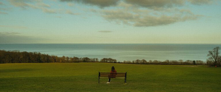 Wide shot of a woman shown from behind, sitting on a bench in front of water