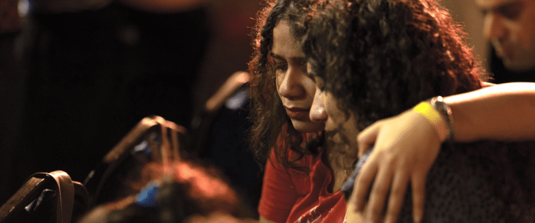 Two women sit closely with their eyes closed as one comforts the other with an arm around her shoulders.