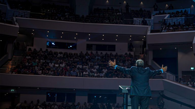 wide shot from the back of the stage of Dr. Youssef, with arms outstretched, speaking to a filled auditorium